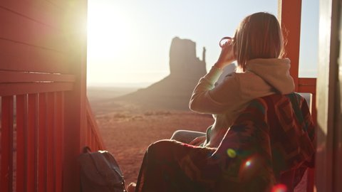 Young woman sitting at hotel cabin terrace looking on cinematic nature landscape in sunrise light. Happy tourist enjoying beautiful morning view and dreaming at trip. Contemplating Monument valley 4K