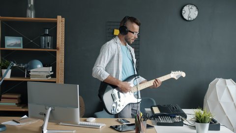 Slow motion portrait of attractive man performer playing guitar and keyboard recording music at home then using computer. Youth culture and technology concept.