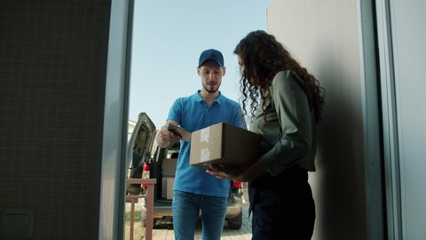 Female customer is opening house front door taking cardboard box from delivery worker signing paper getting order. People and postal service concept.