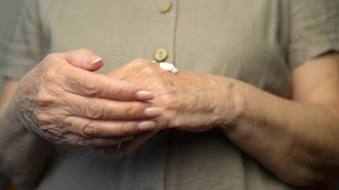 Close-up female hands of senior woman using anti-wrinkle moisturizer, skin care. Mature woman applying cream to her wrinkled hands indoors. Cropped.