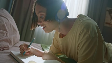 Beautiful smiling girl wearing earbuds lies on bed in bedroom and draws in sketchbook with pencil while listening to music. Creative inspiration concept. Dutch angle slow motion cinematic shot