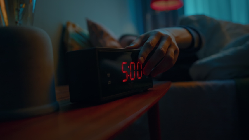 Young female sleeping in bed wakes up at early hour to disable activated digital alarm clock sounding on bedside table. She covers up in blanket and turns away to sleep in. Slow motion cinematic shot