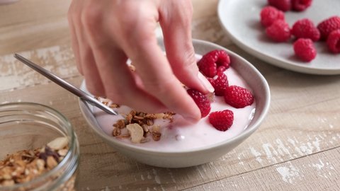 Adding granola and fresh berries into raspberry yogurt bowl. Healthy low calorie high protein and fiber breakfast on wooden table