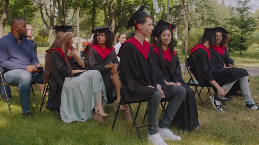 Positive interracial diverse college friends in academic gowns and mortarboards talking and discussing, sitting on chairs on green lawn, preparing for graduation ceremony at university campus. | Shutterstock HD Video #1077002978