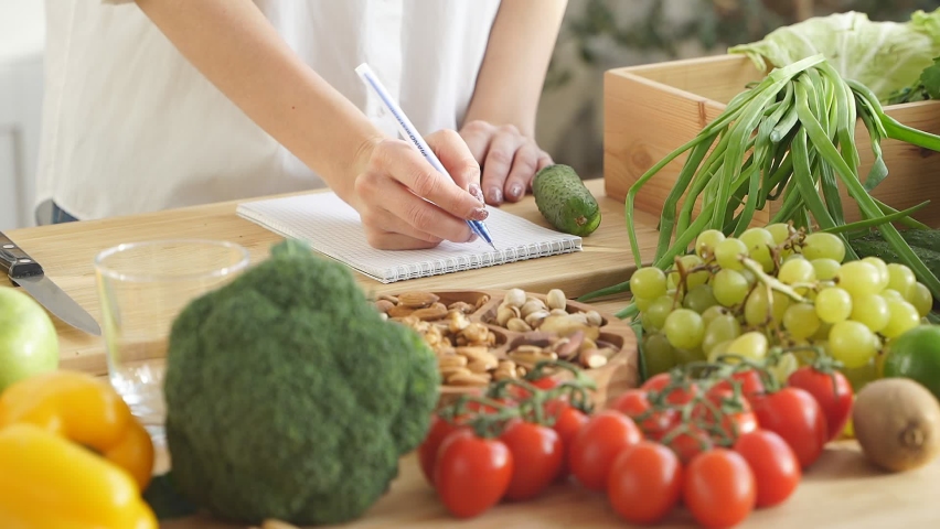Close-up the nutritionist makes notes in his notebook about proper nutrition, there are a lot of vegetables and fruits nearby. In the frame, the hands of a woman without a face. | Shutterstock HD Video #1077006884