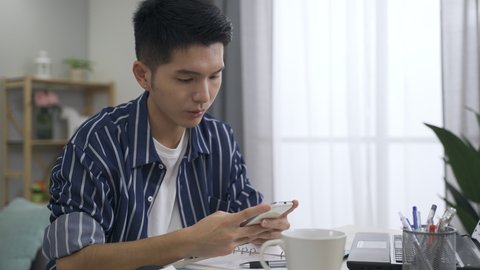 portrait attractive asian guy is mumbling while texting phone message with concentration at the office desk in a bright living room at home.