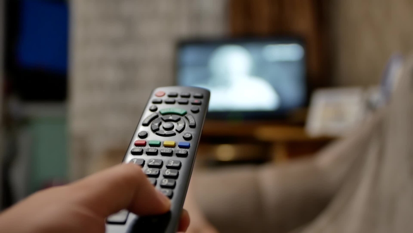 Watching tv close-up hand with remote control switches channels tv on background out of focus | Shutterstock HD Video #1077008321