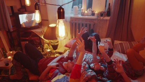 High angle view of company of young friends lying head to head on bed and moving their arms to music while having retro home party in old fashioned room decorated with garland