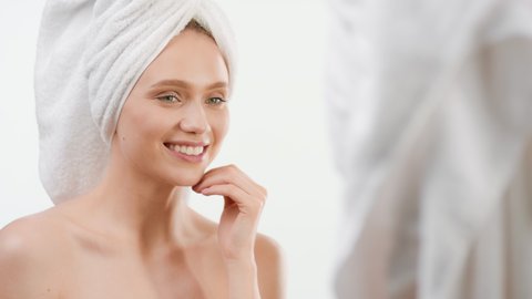 Cute slim Caucasian girl with towel on her head touches her face smiling wide in front of the mirror on white background | Skin rejuvenation concept