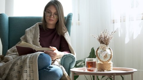 Young woman in eyeglasses reading book, covering cozy plaid, sitting in armchair at home