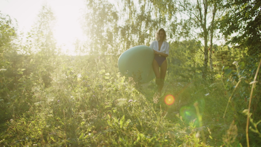 Woman carrying surfing board to lake or river in sunlight. Blonde sportswoman walking in forest with paddleboard. Female coach with surfboard at sunrise. Summer sport and nature, active lifestyle.