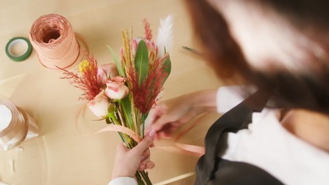 Young florist tying bouquet of roses and dried flowers with ribbon at workplace. Floral artist working in flower shop studio. Floristry creating flower arrangement. Small woman business concept.