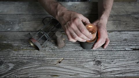 A man lights a wick in a kerosene lamp. A man collects a kerosene lamp at an old wooden table.
