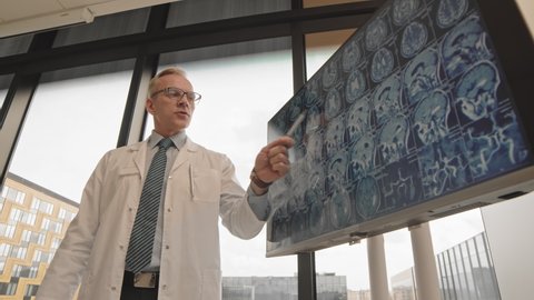 Low-angle medium slowmo of professional male neurologist in lab coat pointing at brain x-ray image on monitor display while making presentation to colleagues