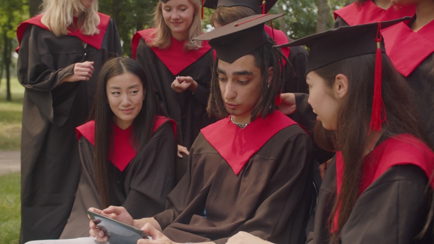 Closeup portrait of group of positive happy diverse multiracial graduates in academic gowns and mortarboards chatting, sharing digital tablet and discussing college years during graduation day. | Shutterstock HD Video #1077055556
