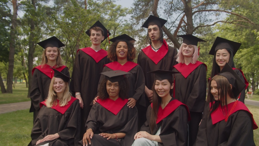 Group of cheerful positive attractive diverse multiethnic graduates in academic gowns and mortarboards celebrating diploma graduation day, expressing carefree mood and happiness outdoors. | Shutterstock HD Video #1077055586