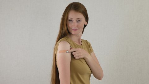Red hair woman patient shows medical plaster on shoulder. Woman demonstrates injection mark. Happy girl satisfied after vaccination with covid-19 virus vaccine. Portrait of girl after vaccination.