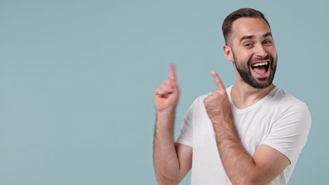 Promoter happy smiling vivid young brunet man 20s wears white t-shirt pointing index finger camera aside on workspace copy space mockup promo commercial area isolated on pastel light blue background