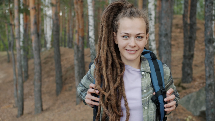 Portrait of young lady with dreadlocks standing outdoors in forest with backpack smiling enjoying wild vacaction. Youth and tourism concept.