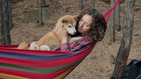 Beautiful young lady is hugging cute shiba inu doggy swinging in hammock relaxing in forest on autumn day. Adventures and recreation concept.