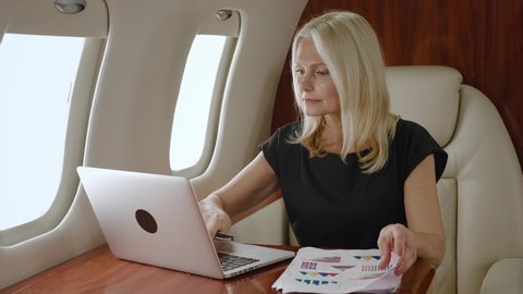 Rich mature businesswoman working with charts, business papers and laptop computer, study the company's growth strategy during a business trip on private jet or first class airplane