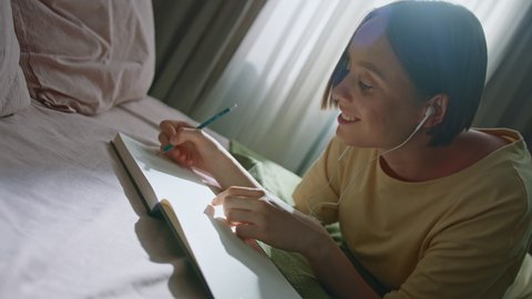 Dutch angle cinematic shot of beautiful smiling girl lying on bed in bedroom and drawing in sketchbook with pencil while listening to music. Calm and positive vibes. Slow motion camera rotation