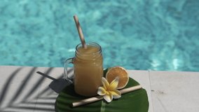 Video footage of glass mason jar with orange juice, bamboo straw, half of fresh orange, yellow frangipani flower, shade from palm tree and bubbling blue swimming pool on background.