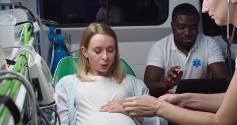 Young pretty Caucasian woman in pain breathing hard and pushing when in labor. Midwife in stethoscope listening heartbeat of baby in ambulance and African American man doc helping.