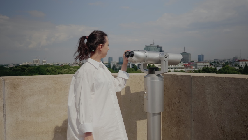 Caucasian tourist looking at panoramic view of metropolis using telescope on sky tower top. Young woman at observation point watching city and nature in metropolitan landscape