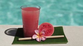 Video footage of glass with pink guava juice, bamboo straw, half of fresh pink guava, tropical flower frangipani and bubbling blue swimming pool on background.