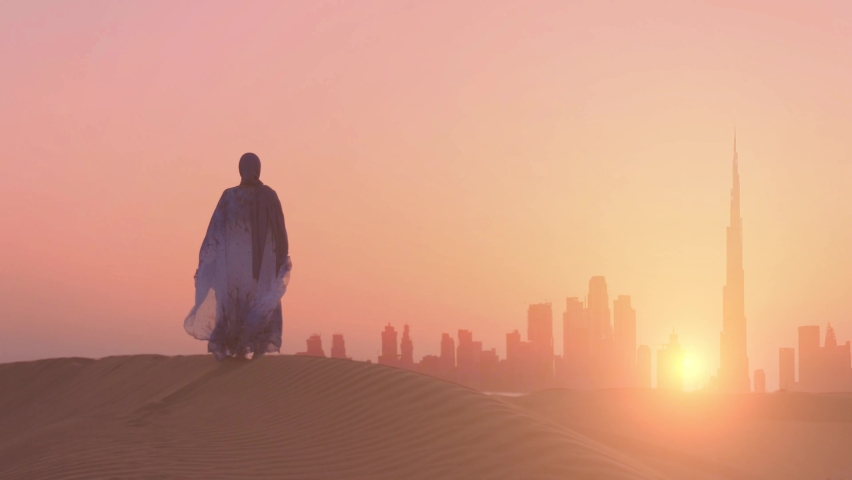 Woman in traditional arab dress stands on the mountains and rises her hand. Dubai city silhouette on the background | Shutterstock HD Video #1077074414