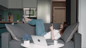 Lazy caucasian woman on headphones lying on the sofa and using smartphone or cell phone in modern room. Tapping, listening to music, scrolling, watching video, content, bloggs.