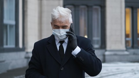 Old businessman, gentleman person in black gloves, coat in coronavirus mask talks on phone. Graying elderly man speaks on smartphone in covid-19 pandemic. Grey haired sick male masked because of covid