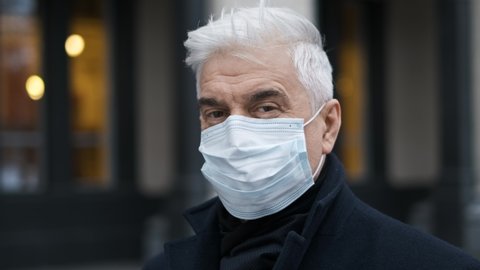 Portrait of old businessman or gentleman person in coat looking at camera. Coronavirus masked people on streets. Gray elderly man walks in covid-19 pandemic. Grey haired male in mask because of covid.