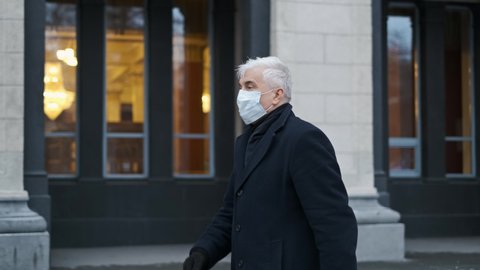 Old businessman or gentleman person in black coat, coronavirus mask on city streets. Graying elderly man walks during covid-19 pandemic with covered face. Grey haired sick male masked because of covid