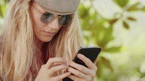 Smiling blonde woman with sunglasses using smartphone, lying relaxing on the hammock in the garden, free time and summer holiday concept to surf the internet or chat with friends using social media