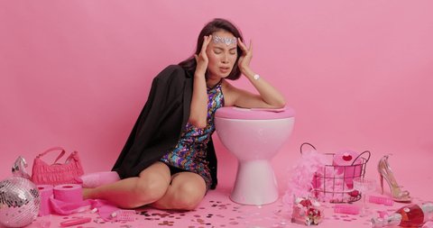 Young Asian woman suffers from severe headache feels unwell after celebration has hangover leans on toilet bowl takes out pills from bag suffers from nausea and dizziness comes home after party