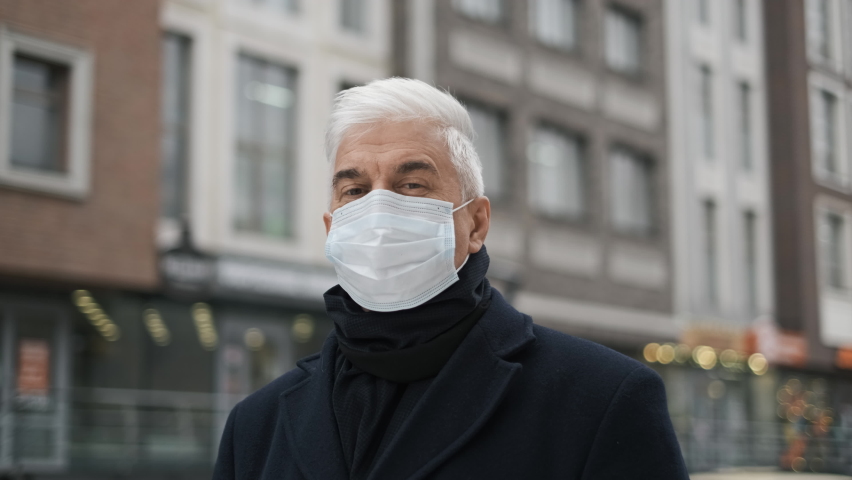 Portrait of old retired businessman, gentleman in coronavirus facemask looks at camera, lockdown restrictions. Mature adult elderly male on city streets. Senior man with graying hair in covid-19 mask.
