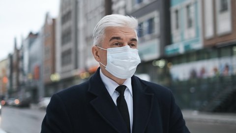 Old retired businessman or gentleman in coronavirus facemask due to lockdown restrictions. Mature adult elderly male in black coat walks on city streets. Senior man with graying hair in covid-19 mask.