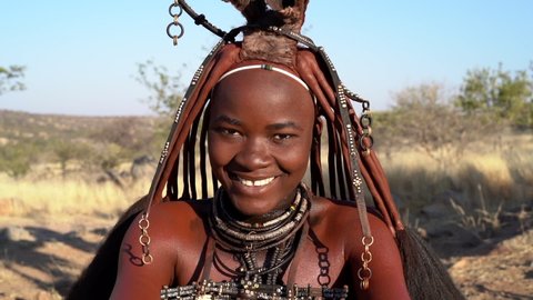Beautiful Himba woman looking at camera and smiling, wearing traditional jewellery and headdress in her village near Kamanjab in northern Namibia, Africa.