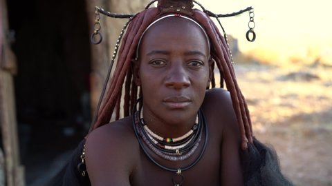 Married Himba woman looking at camera, wearing traditional jewelry and headdress in her village near Kamanjab in northern Namibia, Africa, slow motion shot.