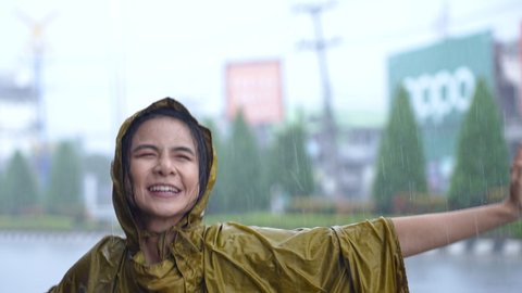young pretty asian girl in yellow raincoat jumping and turning body around on the road side, enjoying the rain falling to her face smiling, rainy season weather pouring rain, feel happy satisfied
