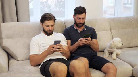 Two gays sit on the couch and use their phones for social networking browsing the internet while show each other photos from smartphone. Next to them sits a beautiful cute dog. LGBT relationship.