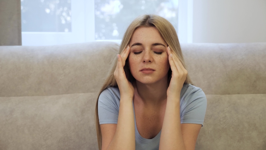 Headache of young woman suffering from migraine at home office touching head sitting on the couch with closed eyes feeling headache. Youth and work concept.