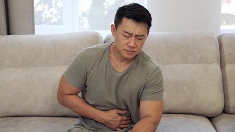 Sick asian man suffering from stomach ache sitting on the couch, holding belly, feeling abdominal or pain in the liver at home. Gastritis, diarrhea and painful periods concept.