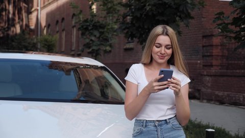 Young Attractive Blonde Woman Standing Near White Car. Using her Modern Mobile Phone. Smiling while reading new Messages. Having Freckles. Beautiful girl in the Old city center. Successful people.
