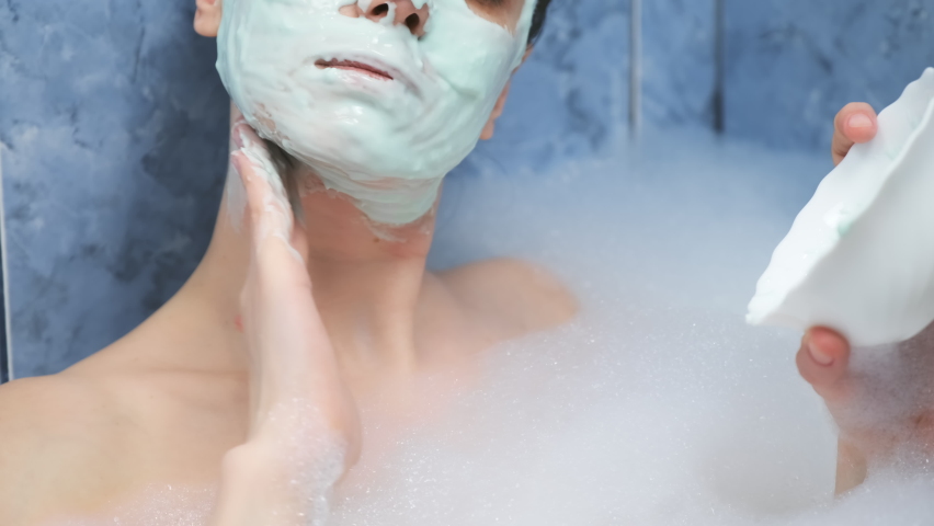 Portrait of young woman is applying alginate mask on face lying in bathroom with foam at home. Spa beauty treatment, anti-aging procedure for face skin. Wellness, skincare and self love concept.