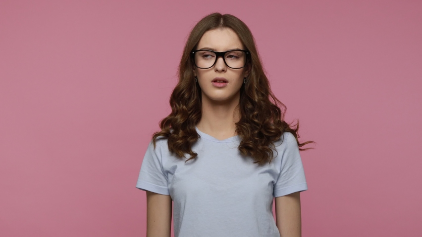 Tired wavy haired female in blue T-shirt takes off eyeglasses and feels exhausted, eye hurt, overworked teenager girl preparing for exam long hours. Indoor studio shot isolated over pink background. Royalty-Free Stock Footage #1077112031