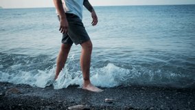 Caucasian man in shorts walks slowly in shallow water of pebble beach. 4K slow motion video of feet in water outside close up. Small sea wave rolls over bare legs. Natural massage for feet and heels.