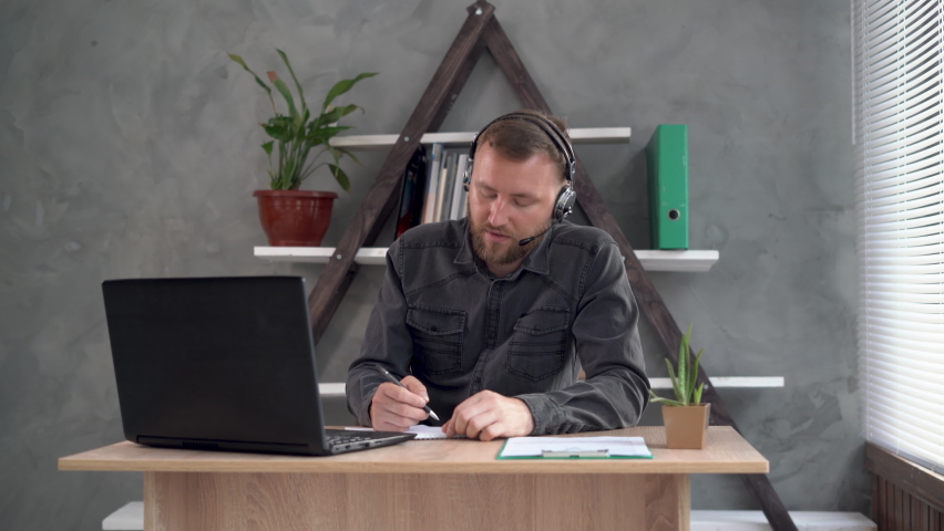 a remote worker, a bearded Caucasian man, sits at the workplace in the office dressed in headphones with a microphone, makes a video call and makes notes in a notebook. Royalty-Free Stock Footage #1077130814